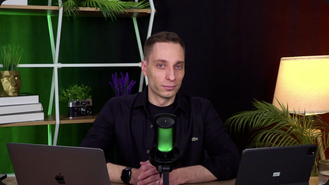 Still from the webinar: Nikita at a microphone between two laptops, a green-lit shelf behind him